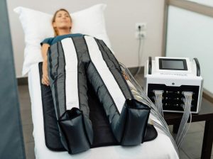 Frau in entspannter Rückenlage trägt eine graue, mit Schläuchen verbundene Lymphdrainage-Hose auf einer Behandlungsliege.