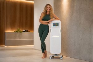 A smiling woman in athletic clothing is leaning against the white Liposana machine with a touchscreen in a modern setting.