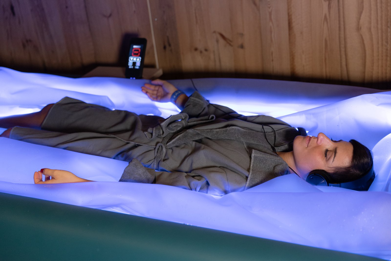 Frau im Bademantel liegt mit Kopfhörern entspannt auf einem blau beleuchteten Dry-Floating-Bett in einem Hotel-Spa
