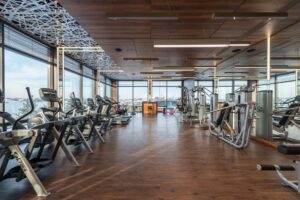 Moderner Fitnessclub mit Cardio-Geräten an der Fensterfront, Holzboden und Kraftbereich im Hintergrund